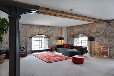 A photograph of a living space with exposed beams, exposed brick wall, a black sofa and a colourful rug on cream carpet.