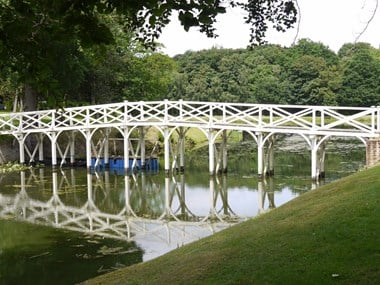 A wooden bridge crossing a river, with a grassy bank in the foreground and mature trees in the background