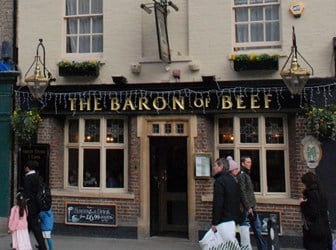 Exterior general view of the Baron of Beef Public House in Cambridge, UK. A black sign with gold lettering reads "THE BARON OF BEEF". In the foreground, people walk past in the street.