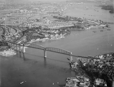 Black and white oblique aerial photograph of a railway bridge spanning a river. The centre of the bridge is dominated by two wide spans carrying massive arched trusses. Boats and barges dot the river. Beyond the bridge are rows of new estate housing and in the far distance are docks harbouring large ships, including two aircraft carriers.