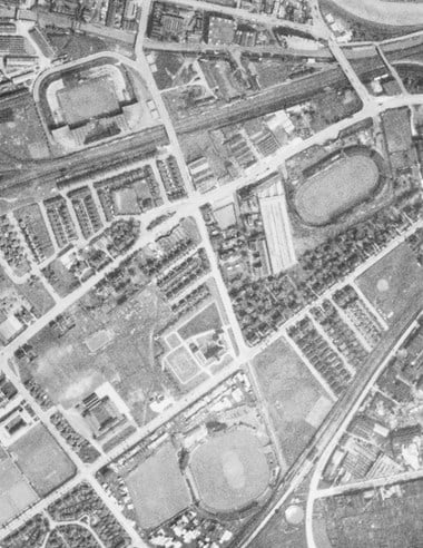 A black and white vertical aerial photograph of part of an urban area, with streets of houses, roads and a railway line. Three sports grounds of similar size and different shapes feature, each with sections of roof over perimeter stands.
