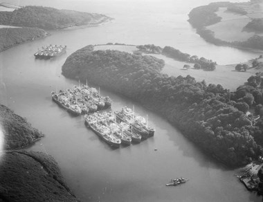 Black and white oblique aerial photograph of a stretch of curving river flanked by wooded hills. The centre of the photograph is dominated by two rows of five closely moored cargo ships. Beyond is a further row of four ships. In the foreground is a small vehicle ferry partway across the river. It carries a variety of cars and vans on its flat, squat deck.