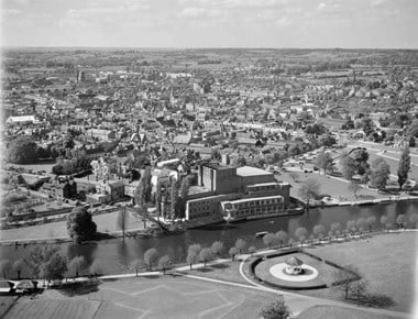 Black and white oblique aerial photograph taken at low altitude. Across the foreground is a stretch of grassy parkland with a bandstand enclosed by a low hedge. Narrow paths and a tree-lined walk follow the line of a river. On the other bank of the river is the Shakespeare Memorial Theatre flanked by grassy parkland, beyond which the town extends towards distant fields, clumps of trees and hills. A narrow stretch of cloudy sky stretches across the top of the photograph.