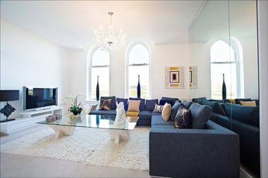 A photograph of a well-lit living room with a blue sofa, white rugs, two large windows, a coffee table and a television. A chandelier-style light hangs overhead.