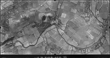 A black and white vertical aerial photograph of a mainly rural landscape. A narrow river winds through the image. Clusters of buildings mark settlements to the top-left, top-right and centre-bottom of the image. Near to the centre of the image are two prominent features, both circular, on of which is encircled by a bank and ditch.