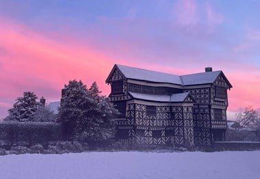 A photograph of a Tudor Manor House set within snowy grounds and pink sunset in the background.