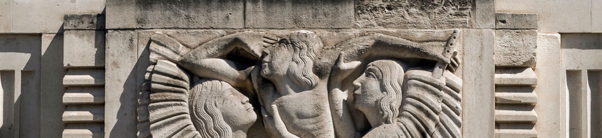 A relief sculpture on a building depicting two winged figures embracing a central figure who  is carrying a flute.