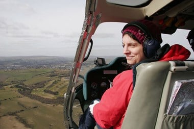 Photograph showing a woman flying a helicopter over a rural landscape. Woman is wearing a knitted hat, red coat and is smiling at the camera.