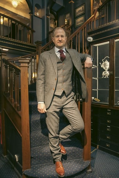 Portrait of a man in a 3-piece suit, standing in a shop, at the bottom of a staircase. 

