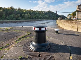 A photograph of a capstan (a metal bollard-like structure) situated on the dockside in Bristol, with a view of the Clifton Suspension Bridge in the background. The capstan is painted black and stands about 1 metre tall. It has a red and white painted band at the top.