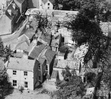 A 1932 black and white aerial image shows The Cedars in North Walsham. The grander house can be seen in the middle surrounded by small and midscale buildings, with trees to the right.
