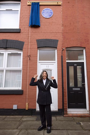 A woman stood on the street outside a red brick terraced house celebrating following the unveiling of a heritage blue plaque. The plaque is in the background mounted on the red brick wall at first floor level.