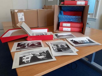Photographs, box files and cardboard boxes on a table. In the foreground are black and white photos of buildings mounted on card. Behind is an open, red box file and closed box files in an open cardboard box.