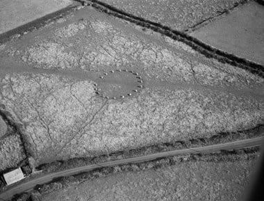 Black and white oblique aerial photograph of a stone circle set in a field and boarded by a track, fields and in the foreground a hedge-lined road. A track runs across the central field and through the ring of stones.