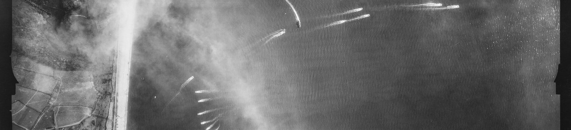 Detail from a black and white vertical aerial photograph, showing ships and their wake on water adjacent to a strip of land. The land is to the left edge of the image and includes a narrow strip of beach next to a patchwork landscape of fields and roads.