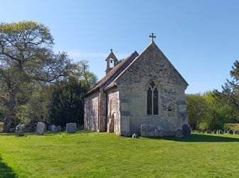 A flint and sandstone chapel stands in a graveyard, with trees beyond. 