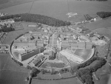 Black and white oblique aerial photograph of a circular prison site surrounded by fields, woodland, residential streets and gardens. Multi-storey accommodation wings and ancilliary buildings radiate from the centre of the prison. A rectangular central area with road and tended lawns extends towards the far side of the prison complex that includes entrance gates and a series of high walls separating the farthest segment of the prison from the rest.