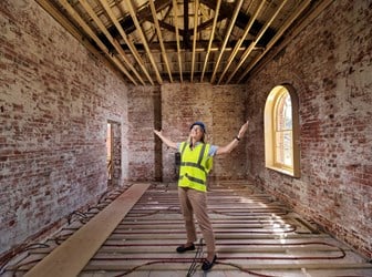 A smiling woman in a high visibility jacket stands in a bare brick room. 