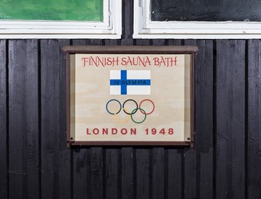 A plaque on the wall of a wooden building with a Finnish flag and the Olympic rings, with text saying "Finnish Sauna Bath, IV Olympia, London 1948". 