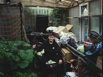 Boy sits in a greenhouse surrounded by plants, boxes and gardening tools.