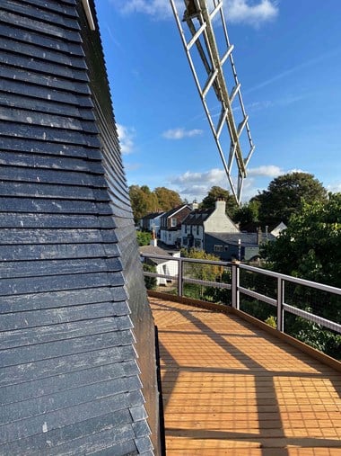 Photograph taken from the second floor decking of Meopham Windmill. I the foreground, you can see part of the main windmill structure, with the newly refurbished oak walkway that wraps around the windmill. In the background you can see houses surrounded by rich greenery and a blue sky.