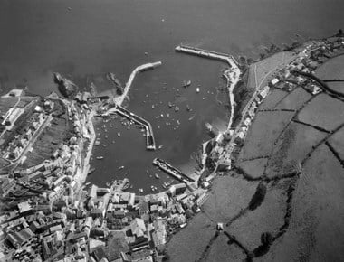 Black and white oblique aerial photograph of a seaside harbour comprising an inner harbour enclosed by two harbour arms and a larger harbour, also enclosed with two harbour arms. Various sized boats dot the harbours. A calm sea is at the top of the photograph and to the right small, enclosed fields on a hillside flank a road that leads to the heart of the fishing port where buildings of various sizes cluster together at the bottom left of the photograph.