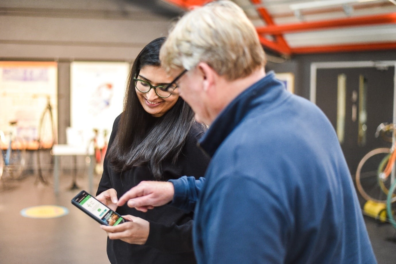 A woman and an older man stand together in a brightly lit indoor space. The woman is holding a smartphone, showing something to the man, who is pointing at the screen