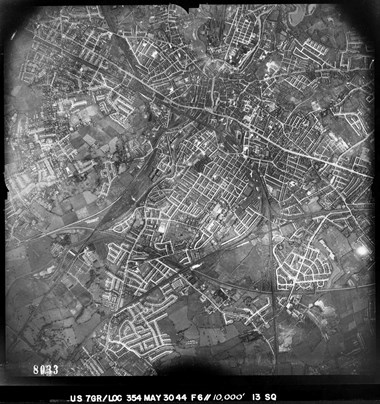 A black and white vertical aerial photograph of an urban area, featuring buildings, roads and railway lines. Patches of fields and open spaces are dotted between built-up areas.