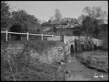 Black and white photograph showing a multi-span, stone bridge over a river. In the background beyond the bridge is a large house. In the left foreground is a section of grassy riverbank sloping up to the bridge approach, which is topped with a wooden fence.
