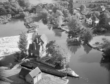Black and white oblique aerial photograph of a mill building standing on a small island of land between a stretch of river, a weir and a lock. The stillness of the river contrasts with the rushing water and white foam of the weir, which rushes leftwards behind the mill. On the opposite side of the river, to the right of the photograph, lawned gardens extend down to the riverside. 