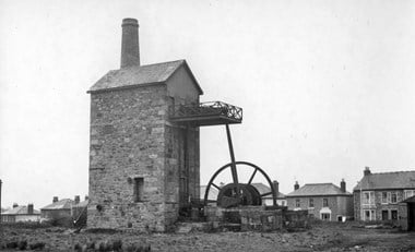 A black-and-white image of a tall stone building with a large metal wheel dwarfing two-storey houses in the background. 