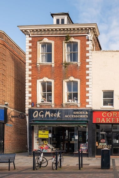 A narrow, red brick three story historic building on a high street. Its ground floor has a modern shop front and signs show it was formerly a shoe shop. It is no longer trading and the building needs repair.