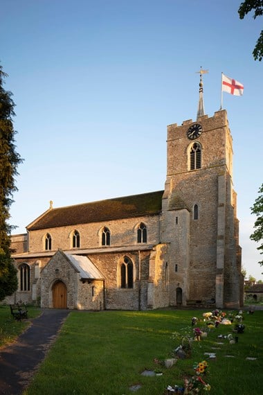 A church flies a St George flag. Fresh flowers have been laid on graves. The afternoon sun streams onto the roof.