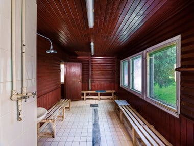 The interior of a wood-clad building, with a tiled floor and drainage channel, a large window overlooking parkland, benches, a shower and sink