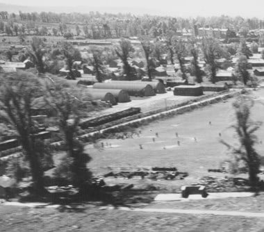 A black and white oblique aerial photograph taken at low level. Figures on a marked pitch in a recreation field play baseball. Beyond the field is a line of huts with curved roofs. In the background are huts dotted amongst trees.
