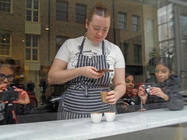 A woman wearing an apron photographed through a cafe window as she photographs coffees whilst also being photographed by children on either side.