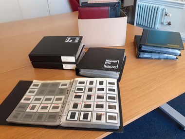 A colour photo of ring-binders on a table. In the foreground is an open binder, showing 35mm slides in clear plastic storage sleeves. In the background is a cardboard box containing four upright binders.
