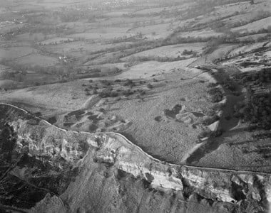 Black and white oblique aerial photograph of a prehistoric earthwork on a hilltop. A curving bank and ditch encloses an area pocked with pits and mounds. In the foreground, the hillside drops dramatically, exposing a steep cliff face topped by a wall. Fields and hedges extend into the distance beyond the enclosure.