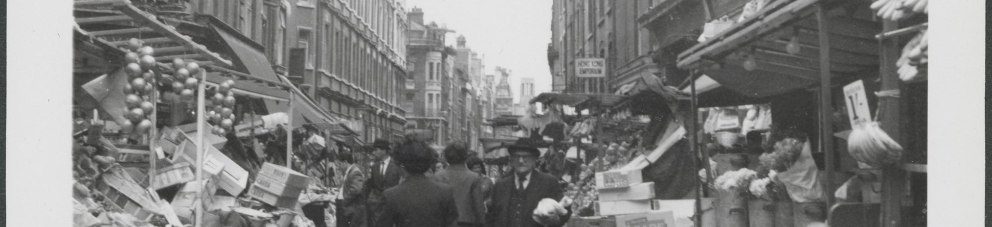 Black and white photograph of an urban street scene featuring people walking in a road flanked by market stalls set out in front of rows of stone-fronted buildings of multiple storeys. 