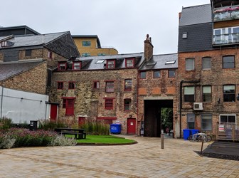 A general view of a courtyard featuring a paved footpath, grassy area with a picnic table, and a listed warehouse building with red doors and windows.