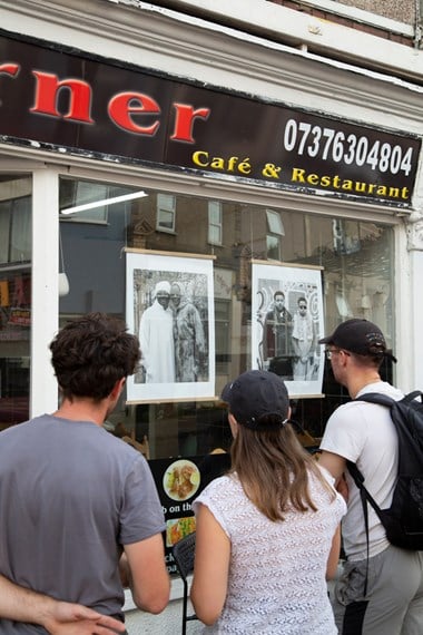 Three people stand in front of a shop window, they are looking at a set of two black and white printed photographs.