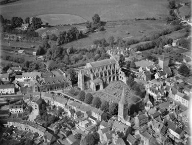 Black and white oblique aerial photograph of a ruined abbey site surrounded by town centre houses, church, market place and roads. The central feature is a surviving abbey church and adjacent graveyard. Extending behind and into the distance are fields, including a cricket pitch.