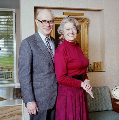 A smartly dressed couple pose for a photograph. The man stands on the left wearing a grey suit. The woman on the right wears a red blouse. They look to be standing in an office.