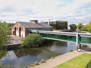 An industrial building on a canal bank, connected to the opposite bank by a long, glass-enclosed walkway.
