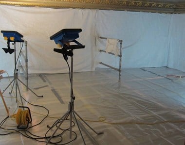 Upright lights stand on a floor covered in plastic sheeting, with white sheets covering the walls.