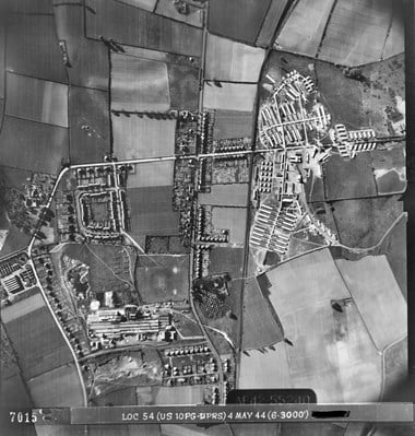 A black and white vertical aerial photograph showing roads, a railway line, fields and buildings, including a factory, houses and an encampment.