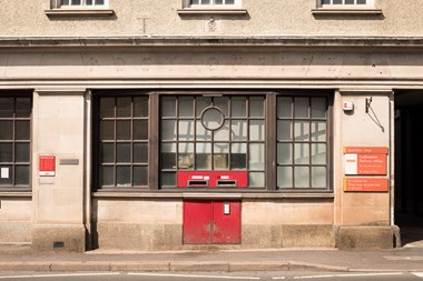 The front of a post office building with a large, multi-paned window above post-boxes which have been set into the masonry.