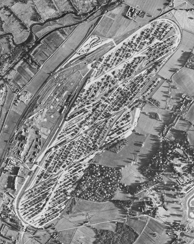 A black and white aerial photograph of an elongated rectangular-shaped site with rounded ends, and filled with rows of objects, possibly containers. The site is bounded by fields and lines of roads, paths and a railway line.