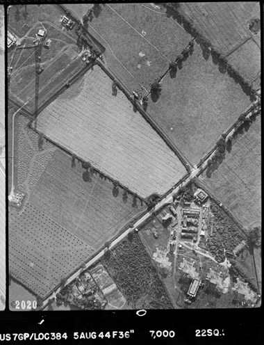 A black and white vertical aerial photograph of irregular fields either side of a road. In the top-left corner is the shadow of a tall tower. In the bottom-right corner are four metal structures adjacent to buildings with camouflaged roofs.