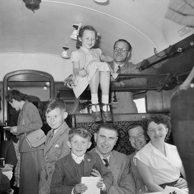 A group of people in a train carriage. Two men sit with a boy and a woman on their knees. Above, a young girl sits on a luggage rack and a man stands on the seat beyond. They all wear suits and dresses, and are smiling. The image is in black and white.